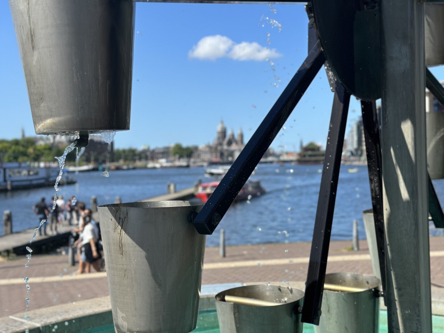 An installation in front of the Technikmuseum. In the foreground a wheel with dripping buckets of water, in the background: the city of Amsterdam