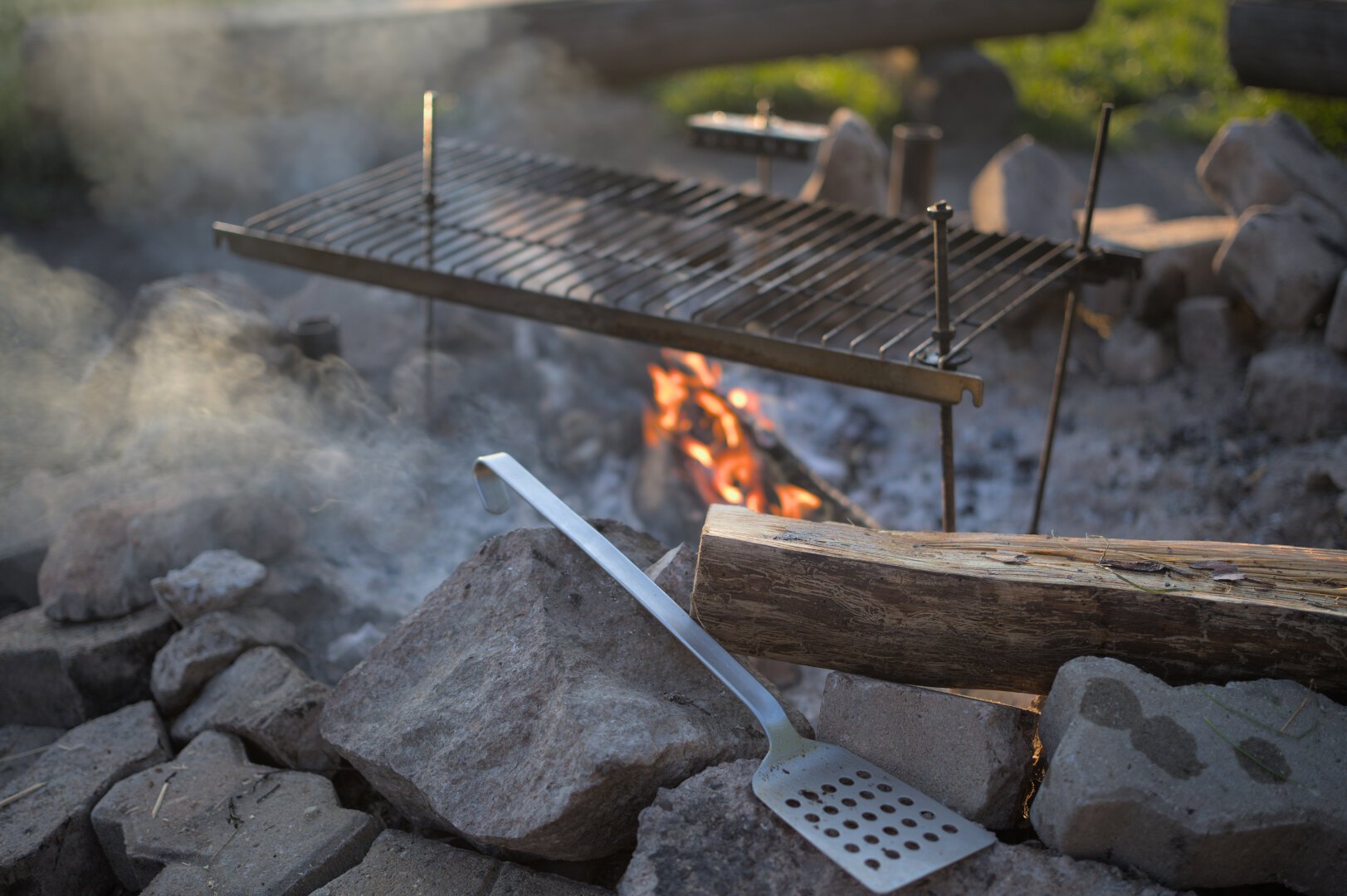 The picture shows a firepit. A small flame is burning visible within it. The focus lies in the foreground where a metal spatula and a wooden log where placed on the stones on edge of the pit.