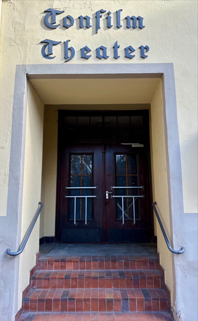 Der Eingang auf dem Bild zeigt eine Tür mit der Aufschrift "Tonfilm Theater". Es handelt sich um ein traditionelles Design mit einer doppelten Holztür. Die Tür ist dunkelbraun und hat oben kleine Fenster. Über der Tür befindet sich eine schlichte Fassade in Cremefarbe mit abgesetztem Grau, die den Namen des Theaters in einer klassischen, leicht gotischen Schriftart präsentiert. Vor der Tür gibt es eine Treppe mit roten Fliesen und ein einfaches Metallgeländer zur Unterstützung. Das Bild vermittelt einen historischen und ruhigen Eindruck.