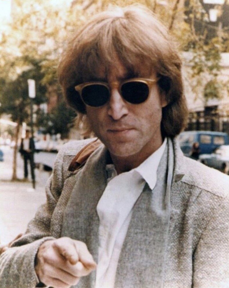 A man (John Lennon) wearing sunglasses and a light gray jacket points toward the camera, with a city street in the background.