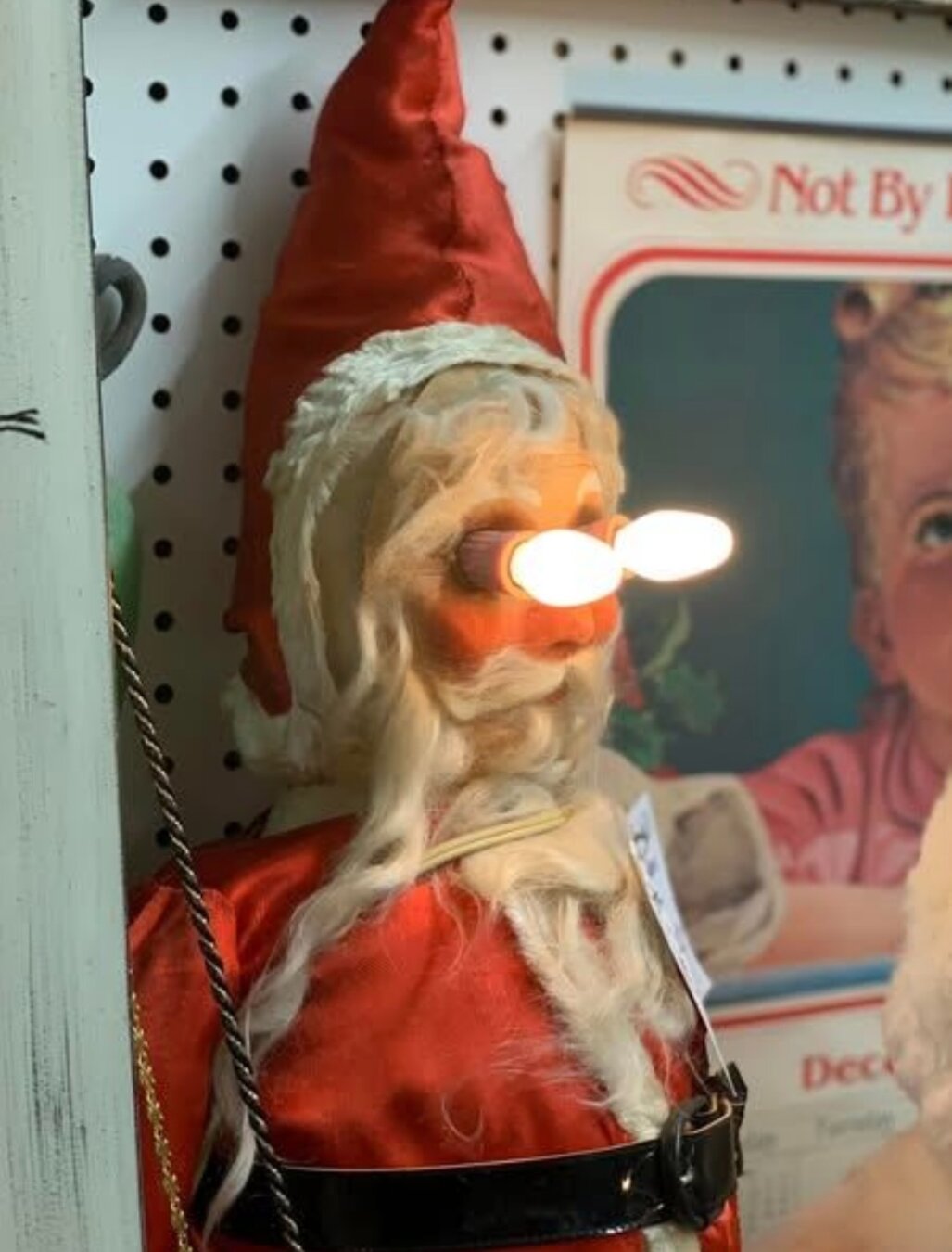 A vintage Santa Claus figure with a red hat and a red outfit. Horrifyingly, white Christmas tree lights have been inserted into his eye sockets.