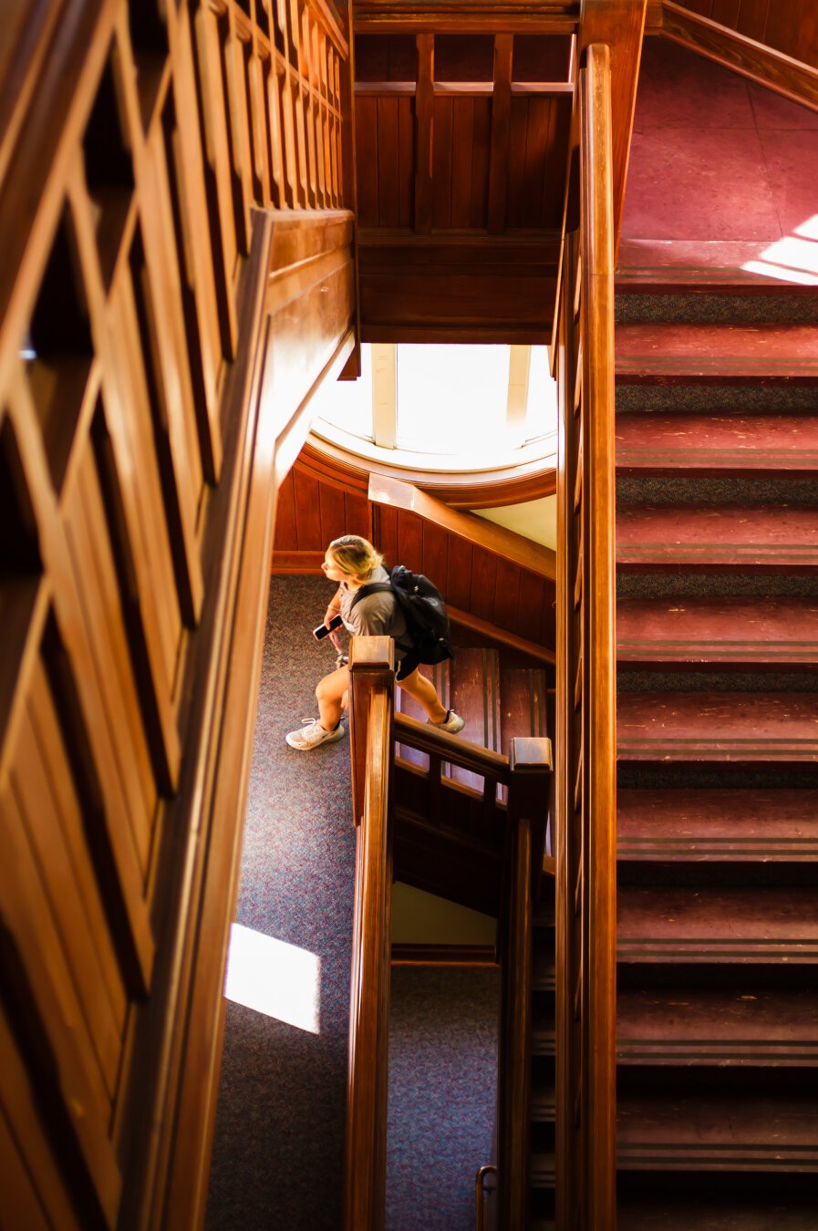 A young woman walking up a stairwell, viewed from a floor above. The stairs are burgundy melamine and the paneling and railing is all oak.