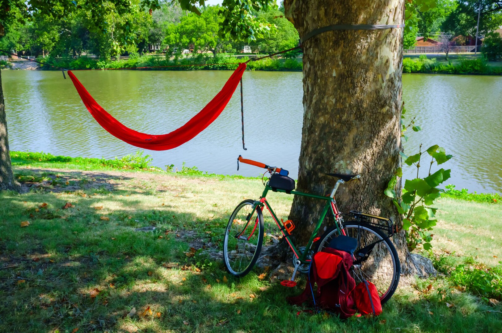 A bicycle parked next to a tree, with an orange hammock hanging from that tree. A city park and river is in the background.