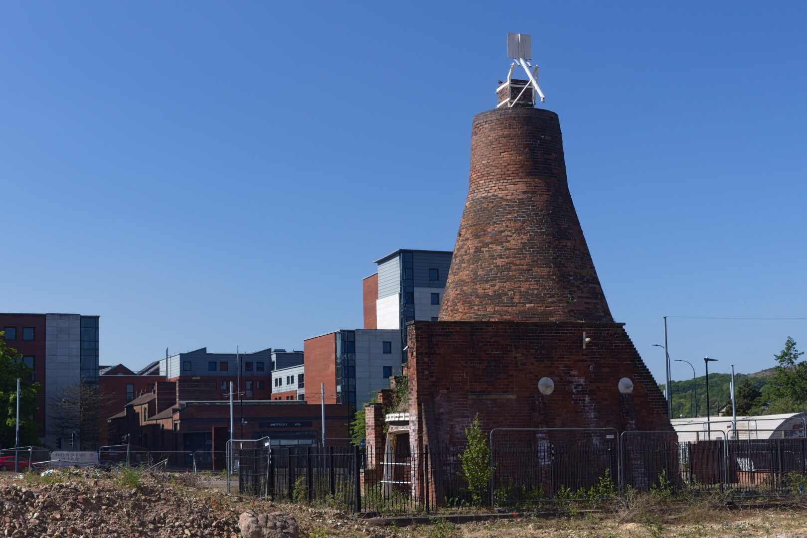 A large, conical brick tower stands prominently in a sunny urban landscape. The tower, once used for cementation in steel production, is dark reddish-brown with weathered bricks and a faded, patchy surface. A modern white metal structure is mounted at its top. Behind it are several mid-rise buildings with red, grey, and white facades, reflecting contemporary architecture. In the foreground lies a rough, dusty area with rubble, overgrown weeds, and metal fencing. The sky is clear and deep blue, casting strong, sharp shadows. The scene contrasts industrial heritage with urban development.