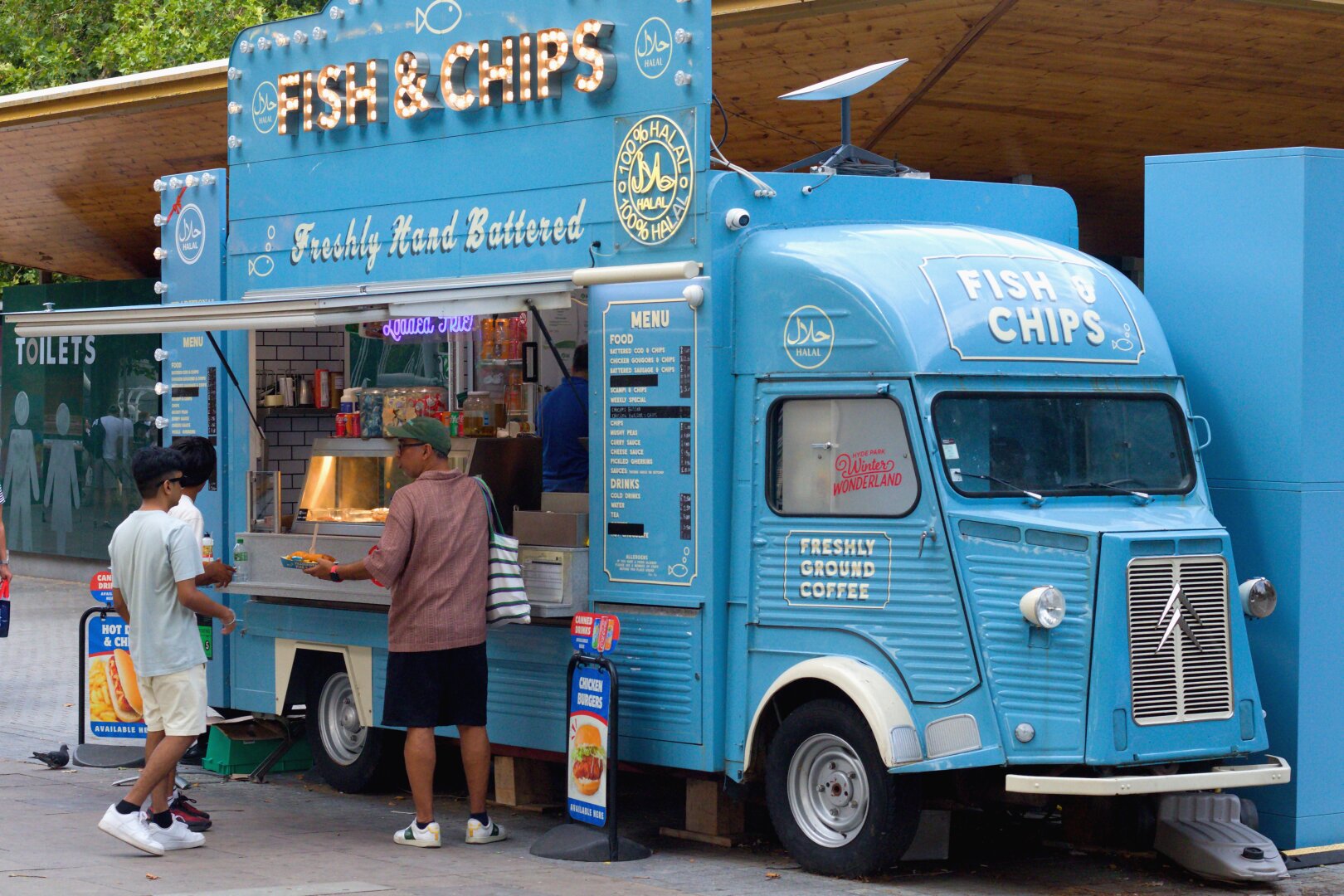 A bright blue vintage-style fish and chips van stands beneath a wooden canopy in a busy London public space. Large illuminated letters spelling “FISH & CHIPS” crown the stall, while menus and hand-painted signs decorate its sides. Three customers wait at the serving hatch: one young man in a pale T-shirt, another partially hidden beside him, and a man in a pink shirt carrying a striped tote bag as he receives his order