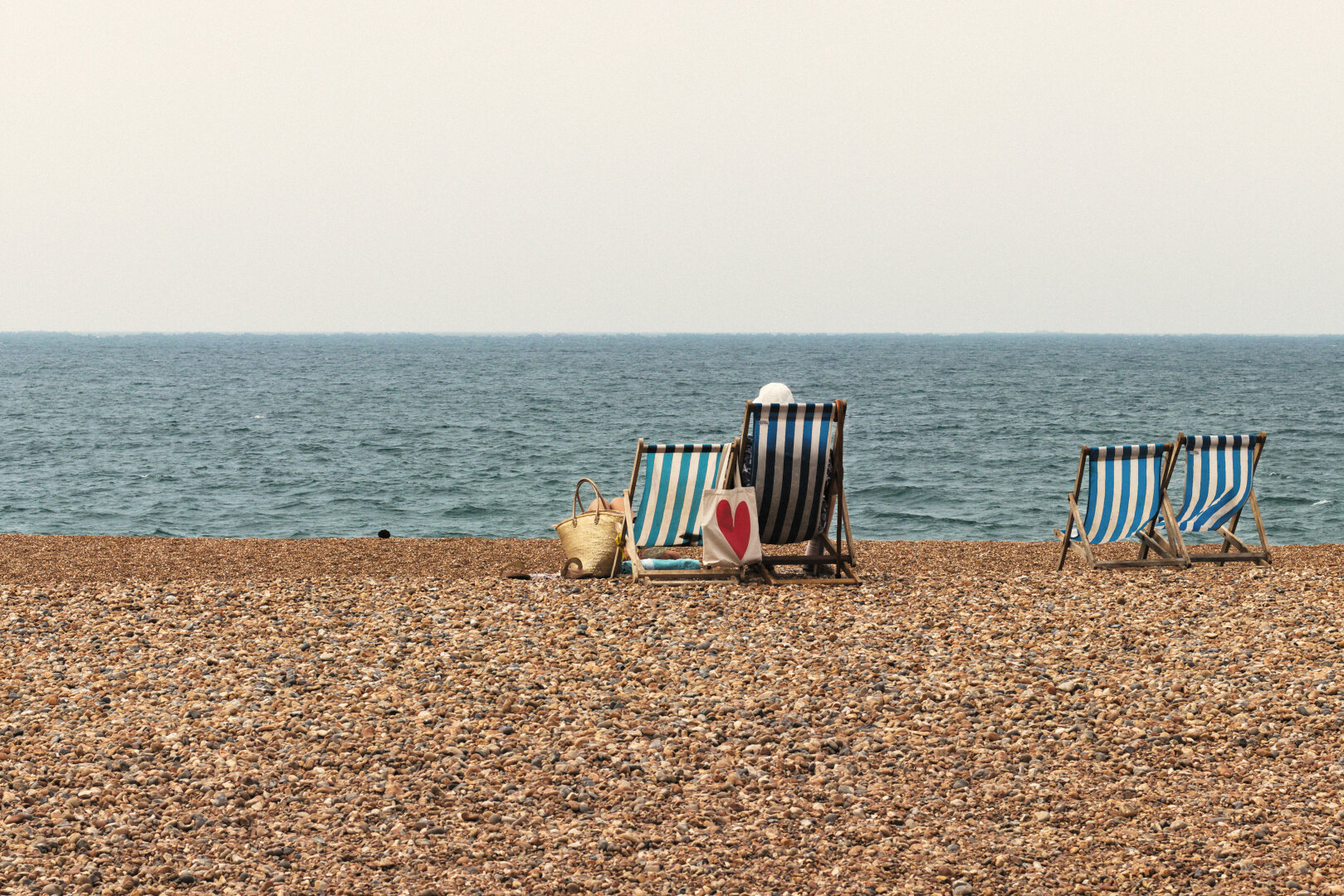 A wide pebble beach stretches across the foreground, leading to a calm blue-grey sea beneath a pale, hazy sky. Near the centre, two traditional blue-and-white striped deckchairs face the horizon. One is occupied by a person in a white sunhat, while another person reclines beside them, mostly hidden from view. Between the chairs sits a cream tote bag with a large red heart, alongside a woven straw beach bag. To the right, another pair of empty deckchairs stands alone, separated by open space.