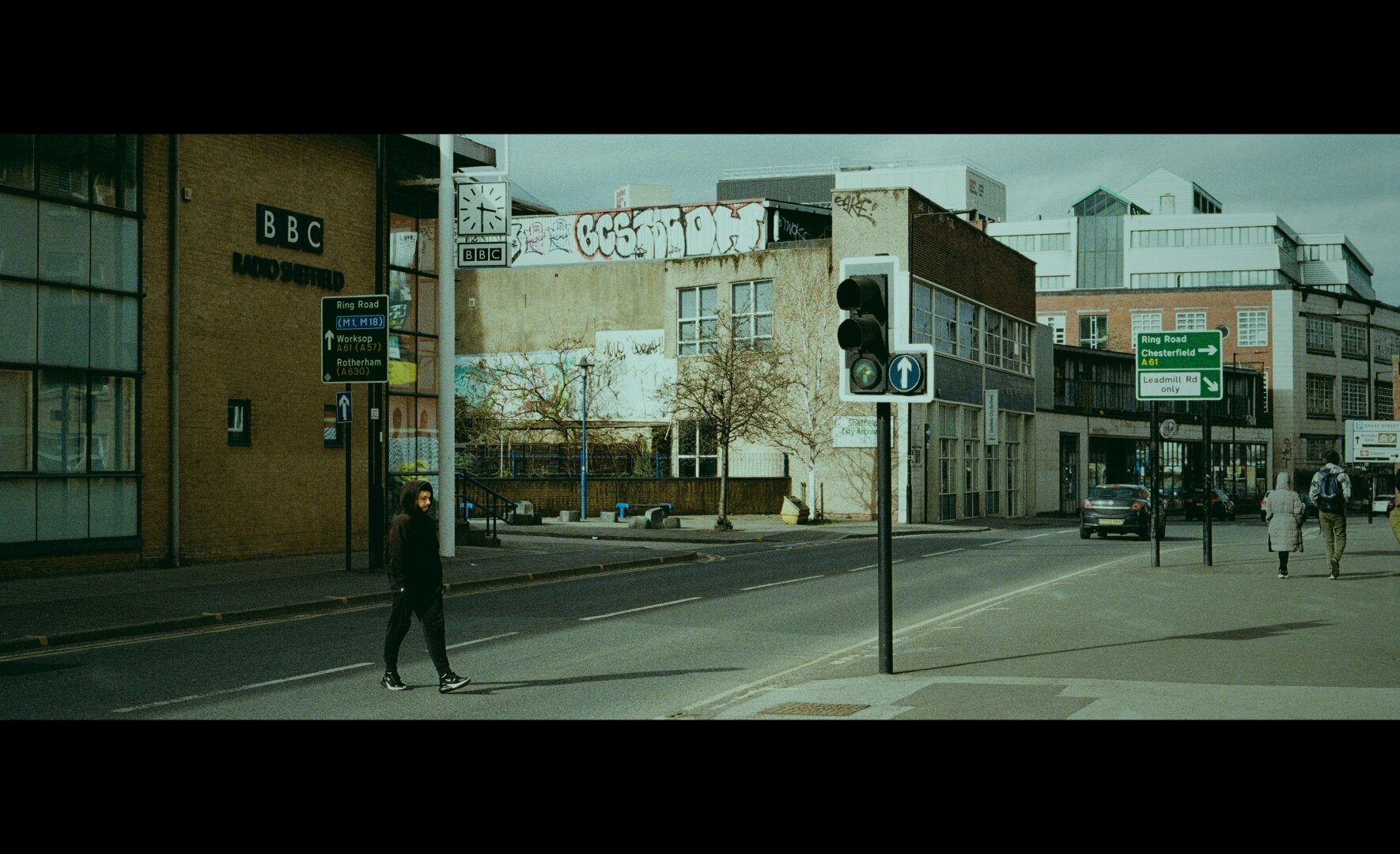 A young person in dark clothing crosses a quiet city street beneath a pale, overcast sky. To the left, a brick building marked "BBC Radio Sheffield" stands beside a street sign and a tall clock. Straight ahead, a faded beige building is covered in graffiti, with bare trees and empty benches nearby. Traffic lights show green, but only a few people are visible—two figures walking away on the right pavement, and a few distant cars. The scene feels still and contemplative, with muted colors and cinematic black bars above and below, adding a sense of framing and introspective distance.