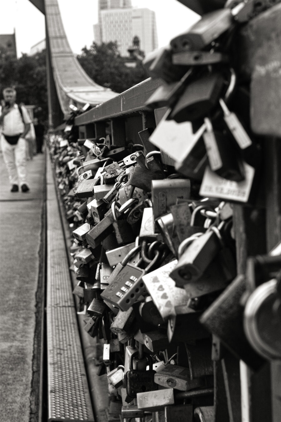 A black-and-white photograph taken on Frankfurt's Eiserner Steg, the famous pedestrian bridge covered with love locks. Hundreds of padlocks crowd the bridge railing on the right, forming a dense mass that stretches diagonally into the distance. The nearest locks are large and softly blurred, while those farther away become smaller and more sharply defined. On the left, a lone pedestrian walks towards the camera, also slightly out of focus. Trees and Frankfurt's buildings rise in the background, softened by the shallow depth of field. The bright sky contrasts with the dark locks and bridge structure, creating a gritty, atmospheric urban scene.