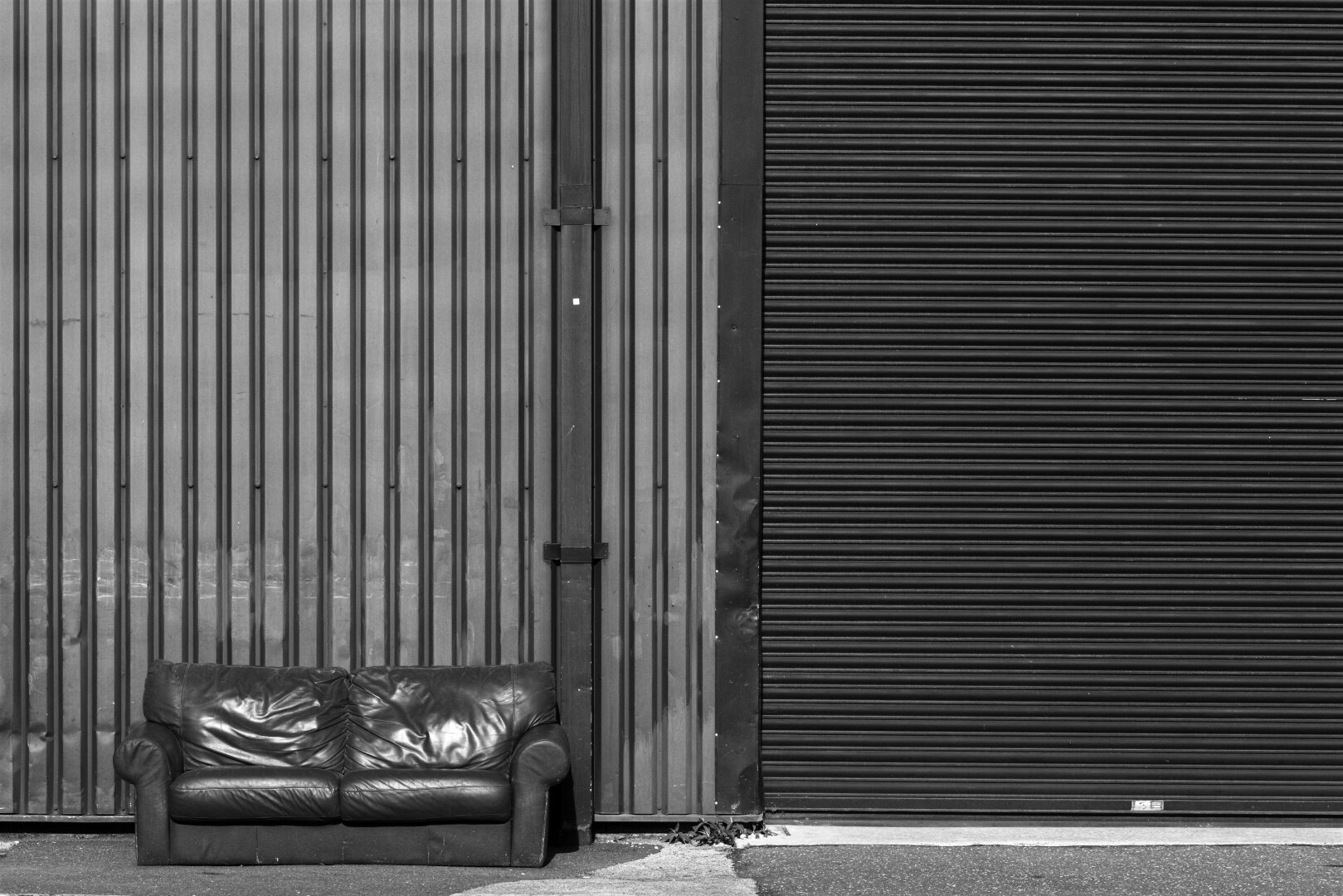 A black and white photograph of a used leather sofa placed outside an industrial building. The sofa is on the lower left side of the picture, on a concrete pavement which stretches through the lower part of the picture. Behind the sofa there’s a tall corrugated metal wall and on the left side of it a large dark roller shutter