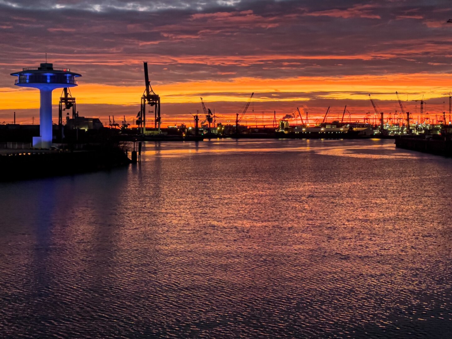 Sonnenuntergang im Hamburger Hafen. IDer rot/orangene Himmel spiegelt sich im Wasser vor den Hafenkränen am Horizont.