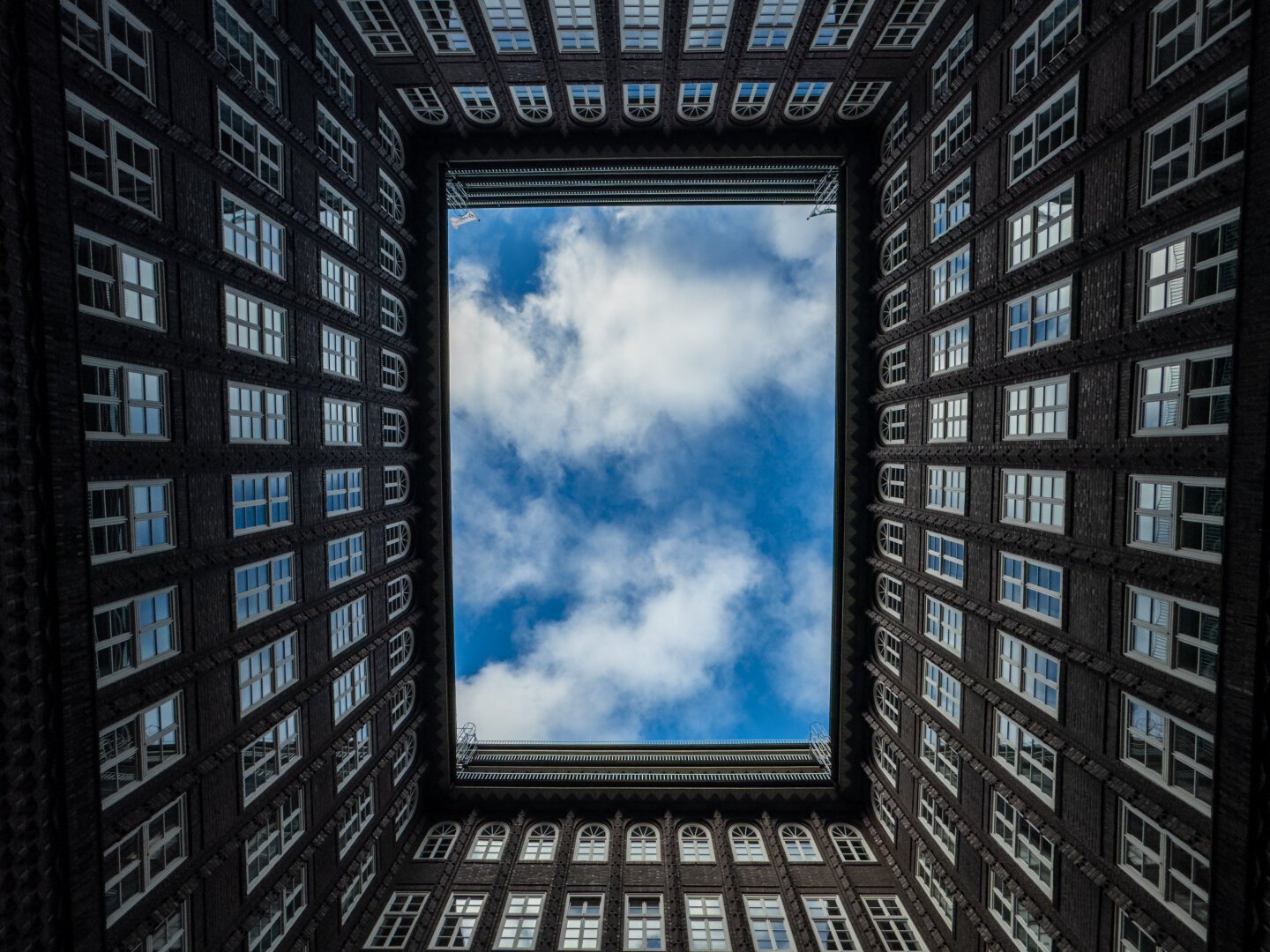 Blick nach oben in einem Innenhof, in der Mitte der Himmel mit ein paar weißen Wolken. Drumherum Fenster in den Innenhof.