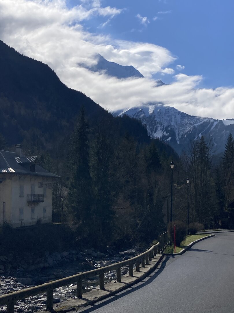 Clouds engulfing huge snow cowered mountains raising up into a sunny blue sky. In the foreground a small road along a rocky mountain river, on the opposide side of the river an abandoned Art Deco style hotel.