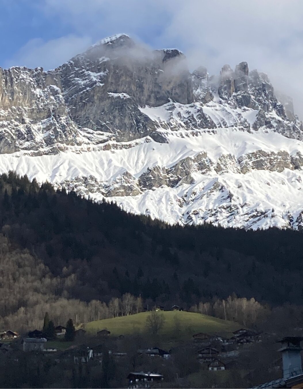 A lush forest with a spot of warm sunshine highlighting two small houses high upon a hill. Behind and above them,tall snow covered cliffs.