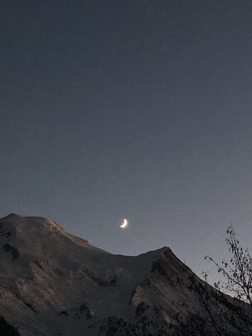 The Mont Blanc mountain and glaciers catching the very last pink rays of sun against a dark sky. The new moon is hanging just above a dip in the mountain ridge.