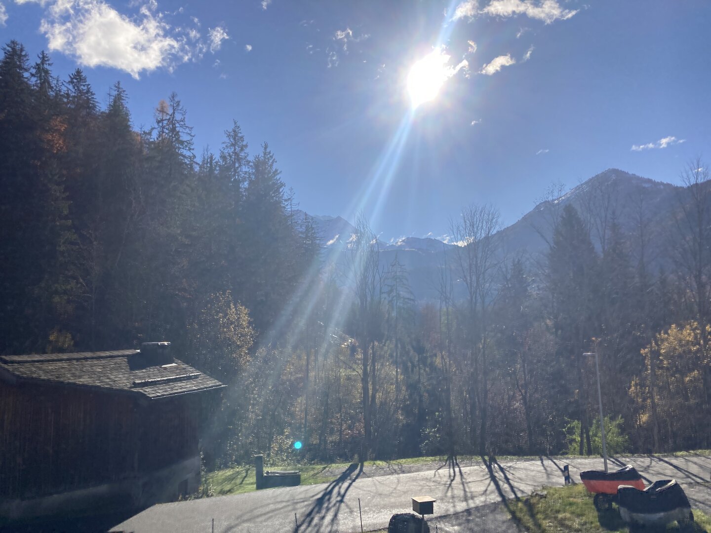 The sun shining down from a blue sky with a couple of small good-weather clouds. Beneath them are some large mountains with snow wind-drifting off the peaks. In front is a forested hill, and a small wooden shed by the bend of a narrow country road.