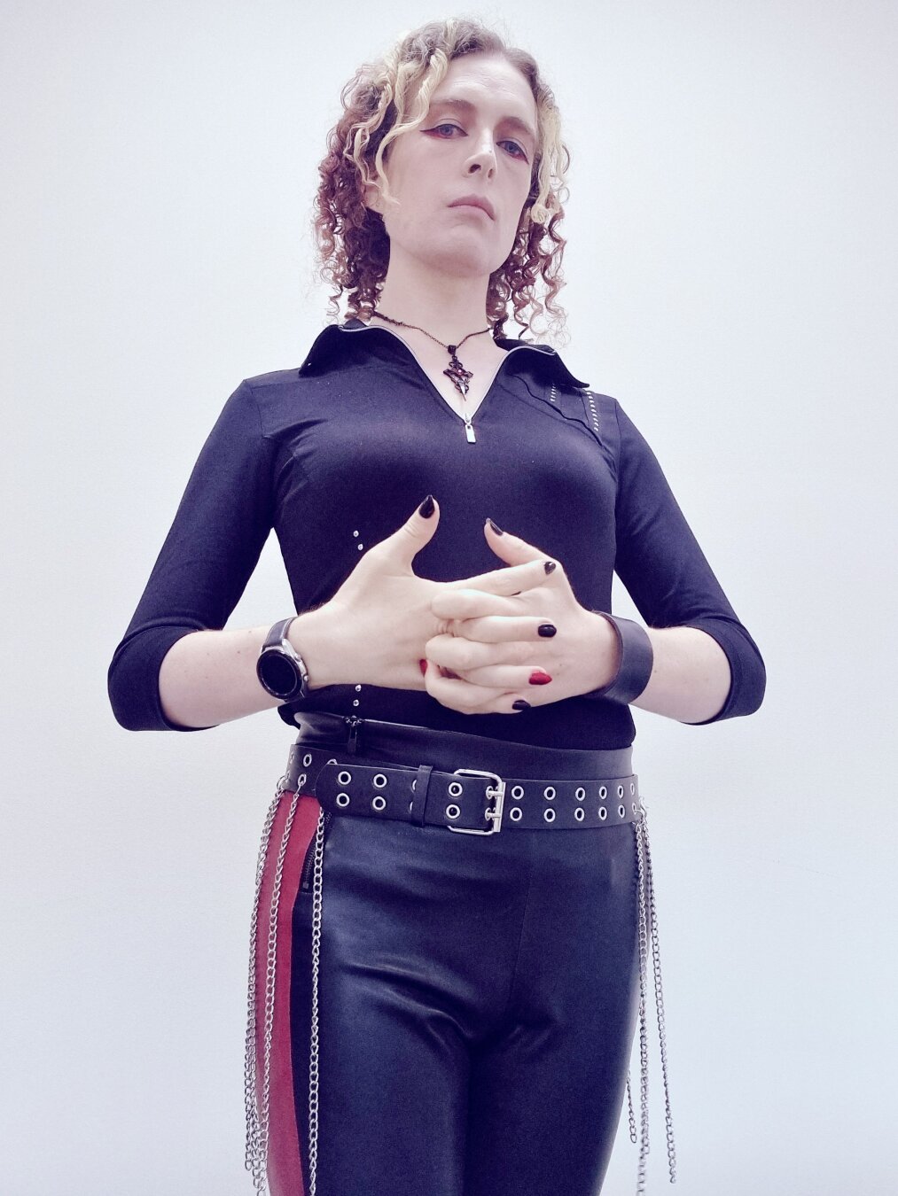 Woman in black/ red leather plants. Studded V neck black top. Chain belt. Gothic necklace. Cracking knuckles