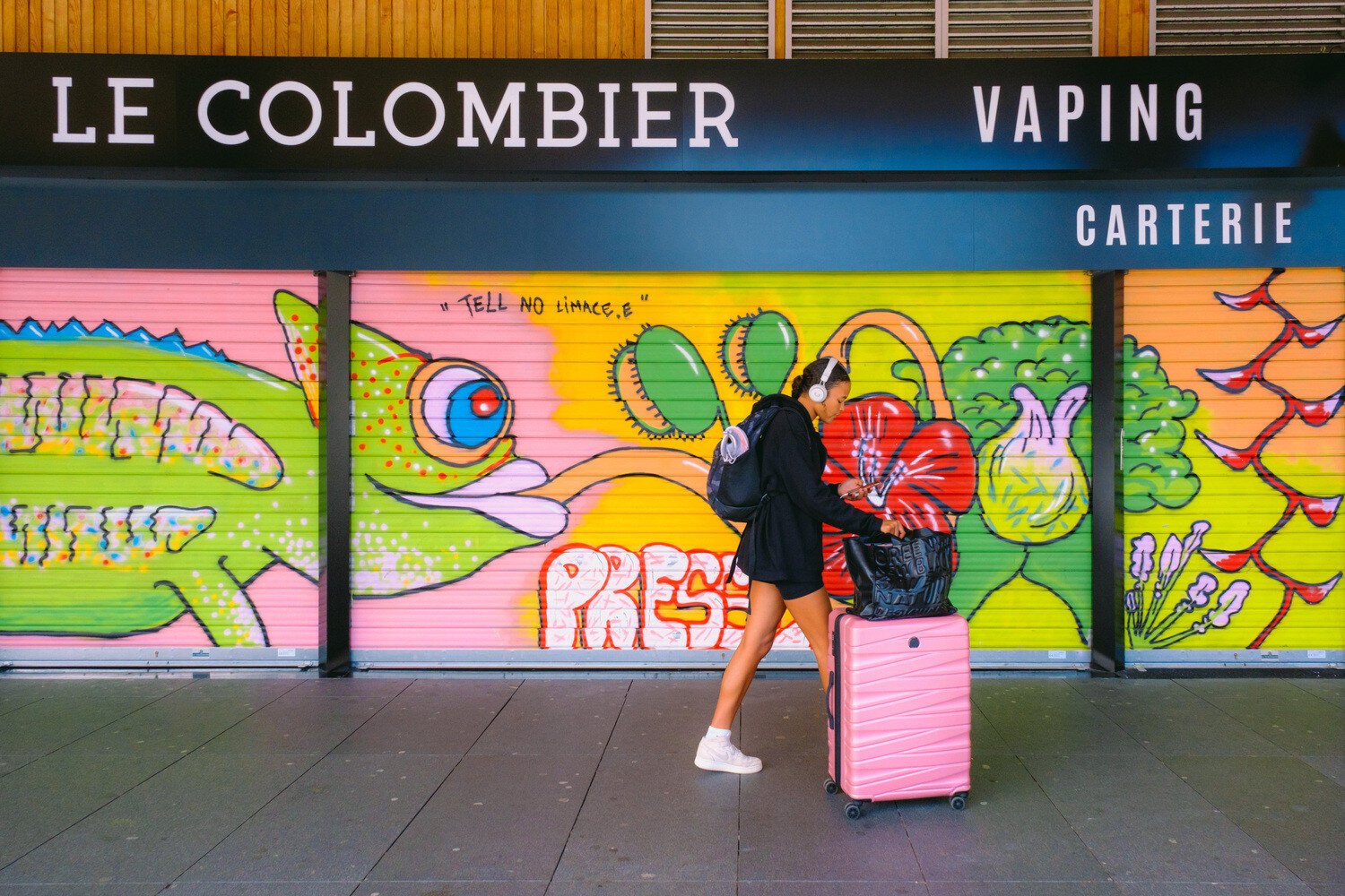 A woman with a pink trolley walks in front of a colorful graffiti. The graffiti contains a chameleon. Its tongue touches the head of the woman.