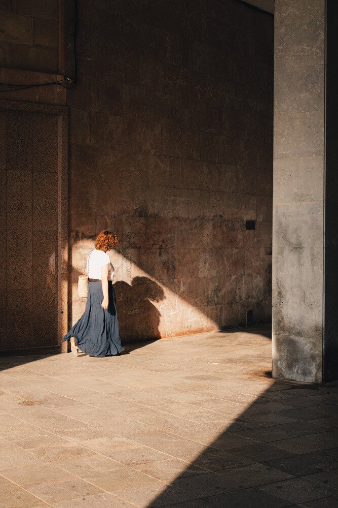 A woman with a long skirt walks through a patch of light. Her shadow is cast on a building wall.