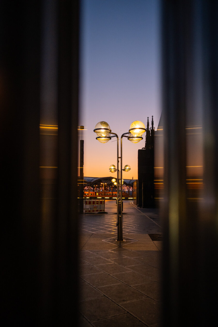 Eine Reihe Laternen stehen hintereinander, im Hintergrund ist der Kölner Hauptbahnhof zu sehen. Der Himmel ist wolkenlos blau, unten sieht man schon die goldenen Farben des Sonnenaufgangs.
Das Foto ist durch metallenen Stützen fotografiert, auf denen sich Reflexionen zeigen Eine Reihe Laternen stehen hintereinander, im Hintergrund ist der Kölner Hauptbahnhof zu sehen. Der Himmel ist wolkenlos blau, unten sieht man schon die goldenen Farben des Sonnenaufgangs.
Das Foto ist durch metallenen Stützen fotografiert, auf denen sich Reflexionen zeigen