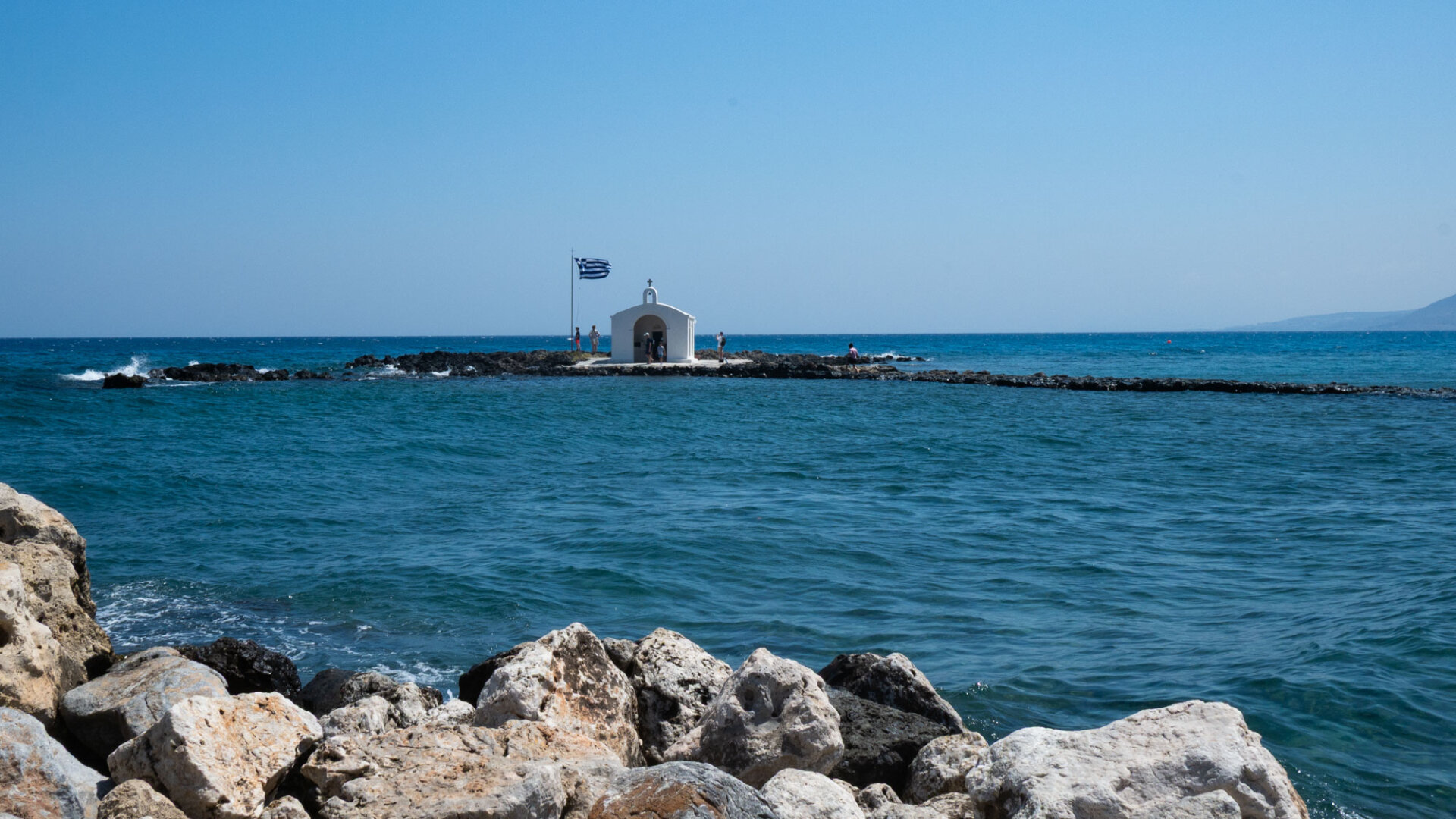 Color photo of a a small white chapel on a rocky outcrop surrounded by the blue sea. Large rocks fill the foreground, while a Greek flag flies beside the chap