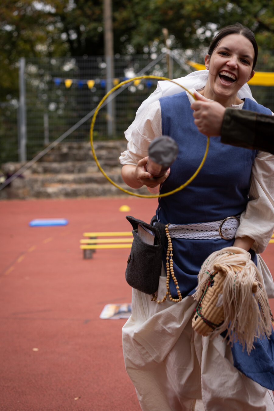 A smiling woman in a blue-and-white costume rides a hobby horse while holding a padded makeshift jousting pole. Another person’s hand holds a medium size hoop in front of her as a jousting target, with playground equipment and trees visible in the background.
