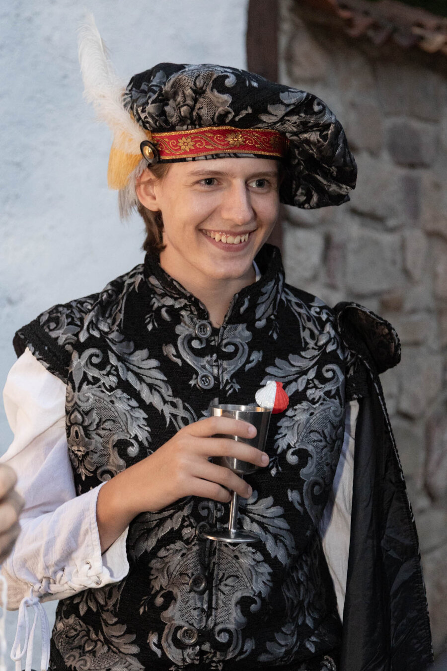 Color photo of a smiling person in an ornate black-and-silver costume with a decorative hat adorned with a feather. They are holding a metal goblet against a stone-and-plaster background.