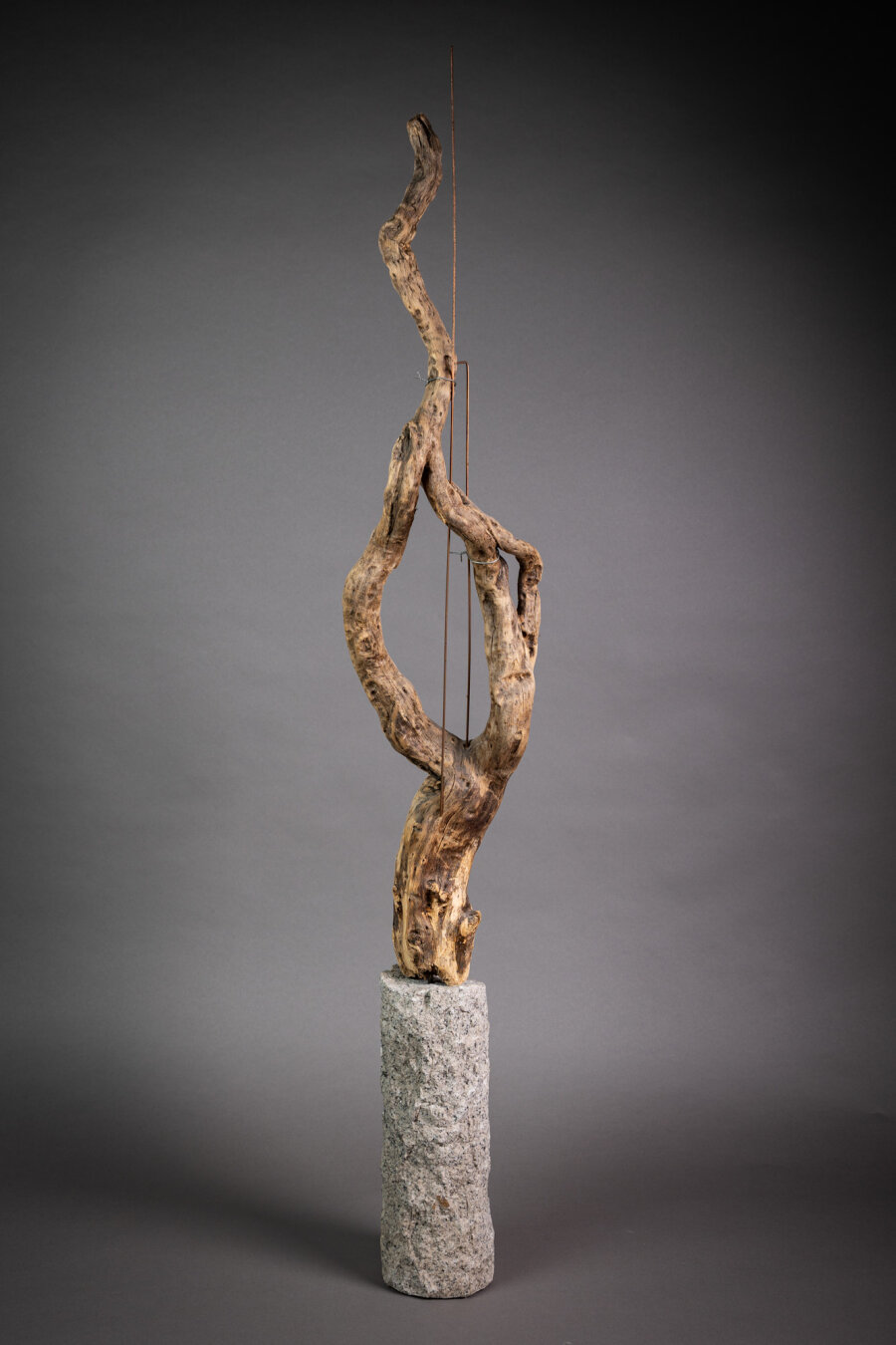 Color photo of a tall, twisted piece of natural wood mounted on a cylindrical stone base. Thin vertical wires run alongside the wood against a gray background.