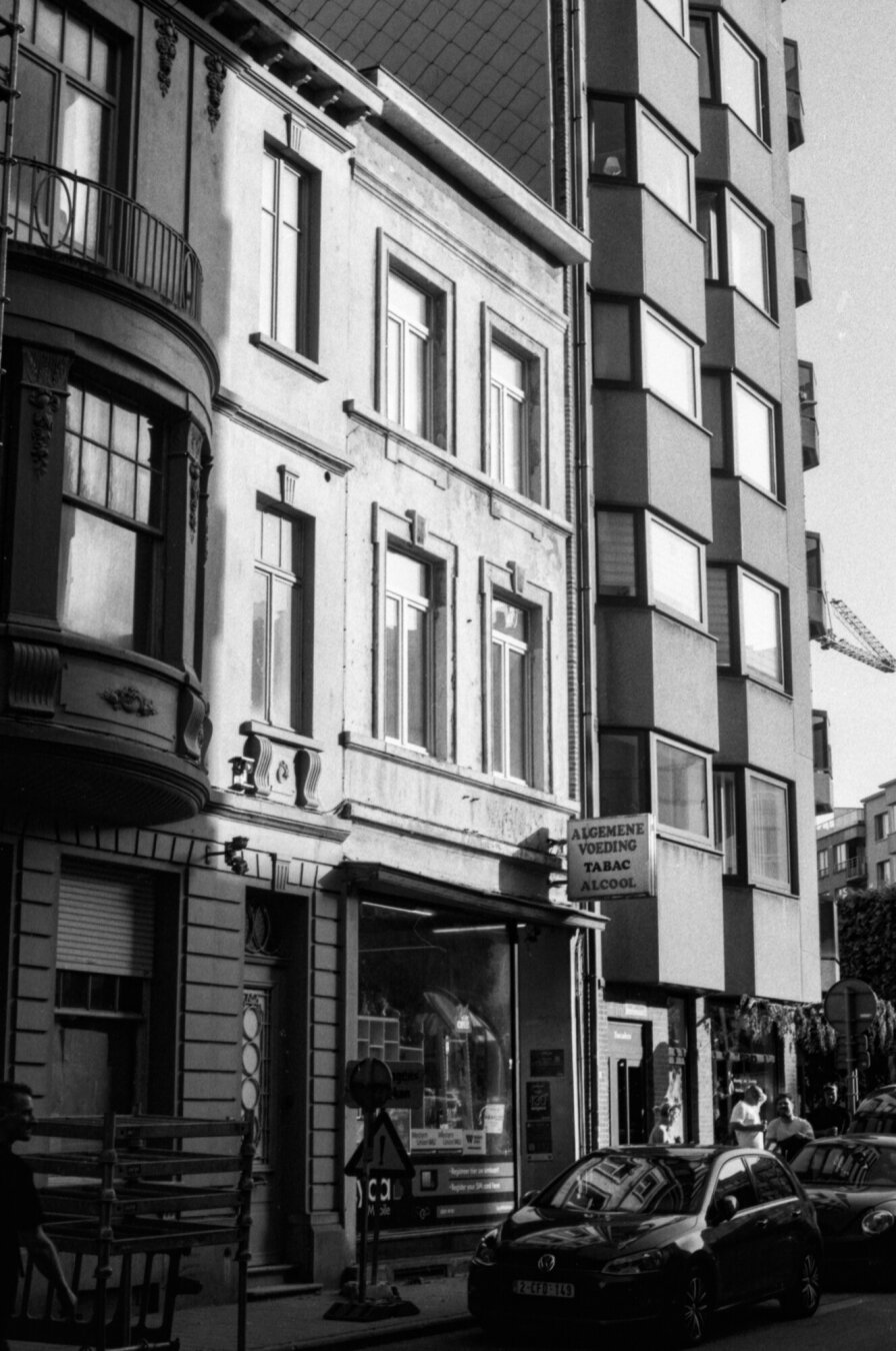 Black-and-white photo of a urban street lined with multi-story buildings, including an older ornate facade beside a modern apartment building with bay windows. Parked cars and storefronts at street level suggest a busy city neighborhood with mixed residential and commercial use.
