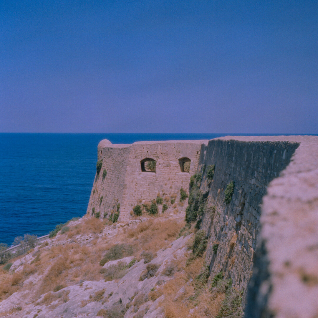 Color photo of a weathered stone fortress wall overlooking a deep blue sea. Dry grasses and vegetation cover the rocky slope beneafth the fortifications.