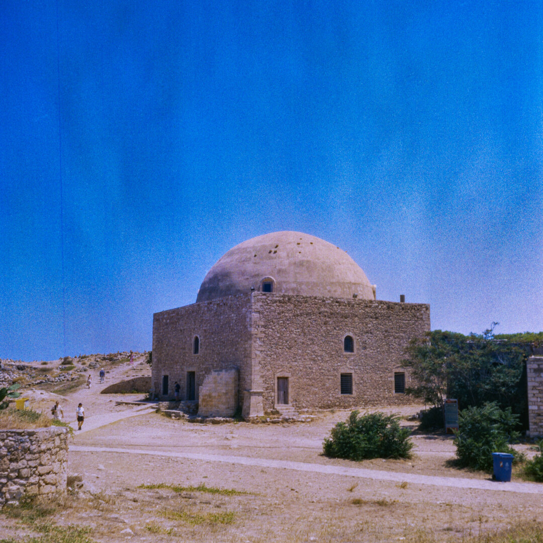 Volor photo of a a rectangular stone building with a large beige domed roof standing in a dry, rocky landscape beneath a bright blue sky. Several people walk along the sandy paths around it, with sparse shrubs, a low stone wall,A low, rectangular stone building with a large beige domed roof stands in a dry, rocky landscape beneath a bright blue sky. Several people walk along the sandy paths around it, with sparse shrubs and a low stone wall nearby.