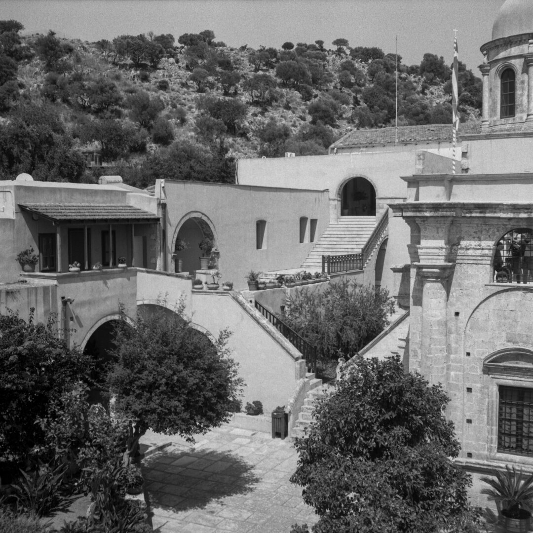 Black and white photo of a sunlit stone courtyard featuring arched doorways, tiled roofs, stairways, and lush Mediterranean plants, creating a serene historic atmosphere. Behind the buildings, a rugged, tree-covered hillside rises under a clear sky, while a domed stone structure frames the right side of the scene.
