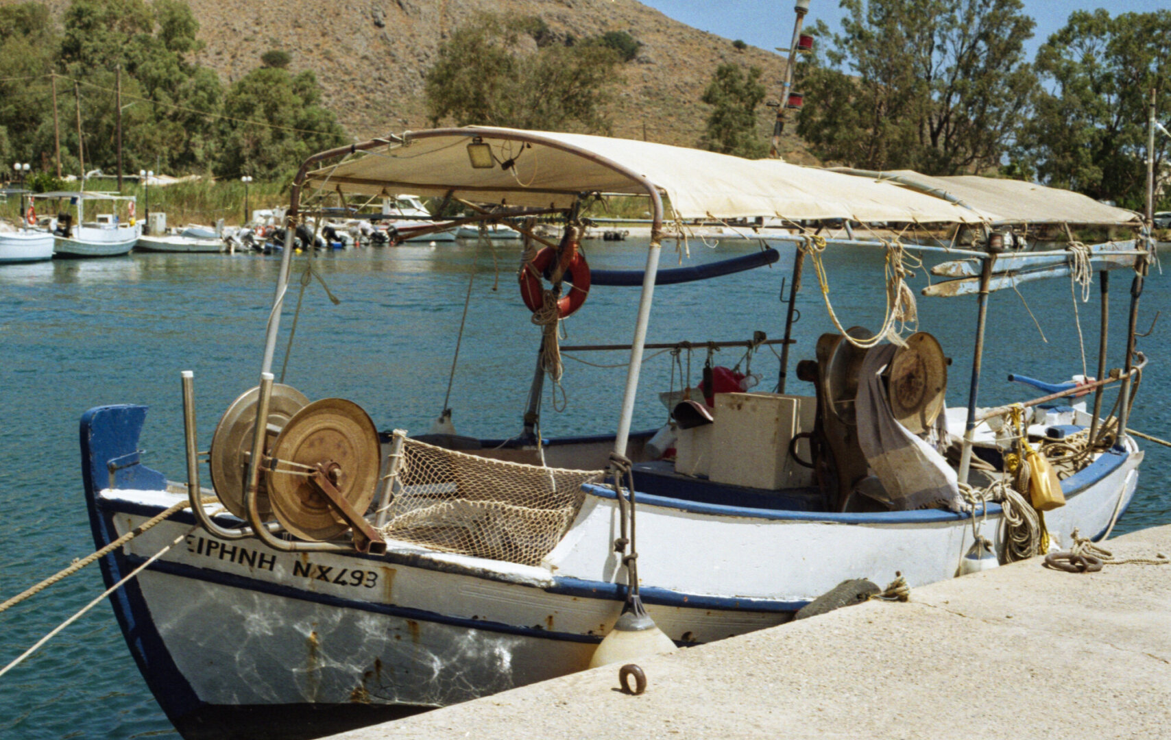 Color photo of a small white fishing boat moored beside a concrete dock, with a canopy, fishing gear, ropes, nets, and a life ring on board.
Behind it are calm blue-green water, several other boats, tall trees, and a dry, rocky hillside under a clear blue sky.