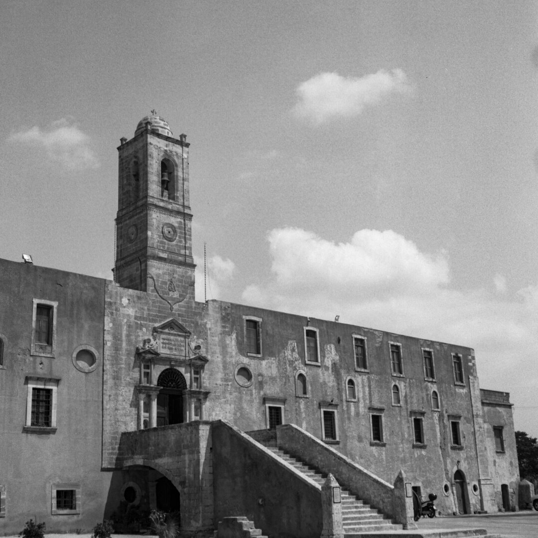 Black and white photo of a weathered historic stone building with a tall bell tower and ornate arched entrance. A broad staircase rises to the doorway. Puffy clouds fill the sky above the old façade.