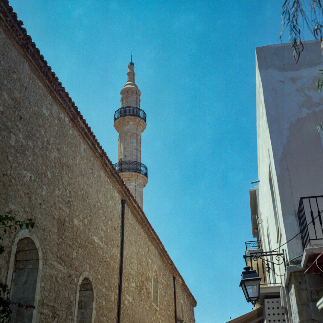 Color photo of a tall, ornate stone minaret above an old weathered wall beneath a brilliant blue sky. Narrow historic buildings and wrought-iron balconies line the sunlit street