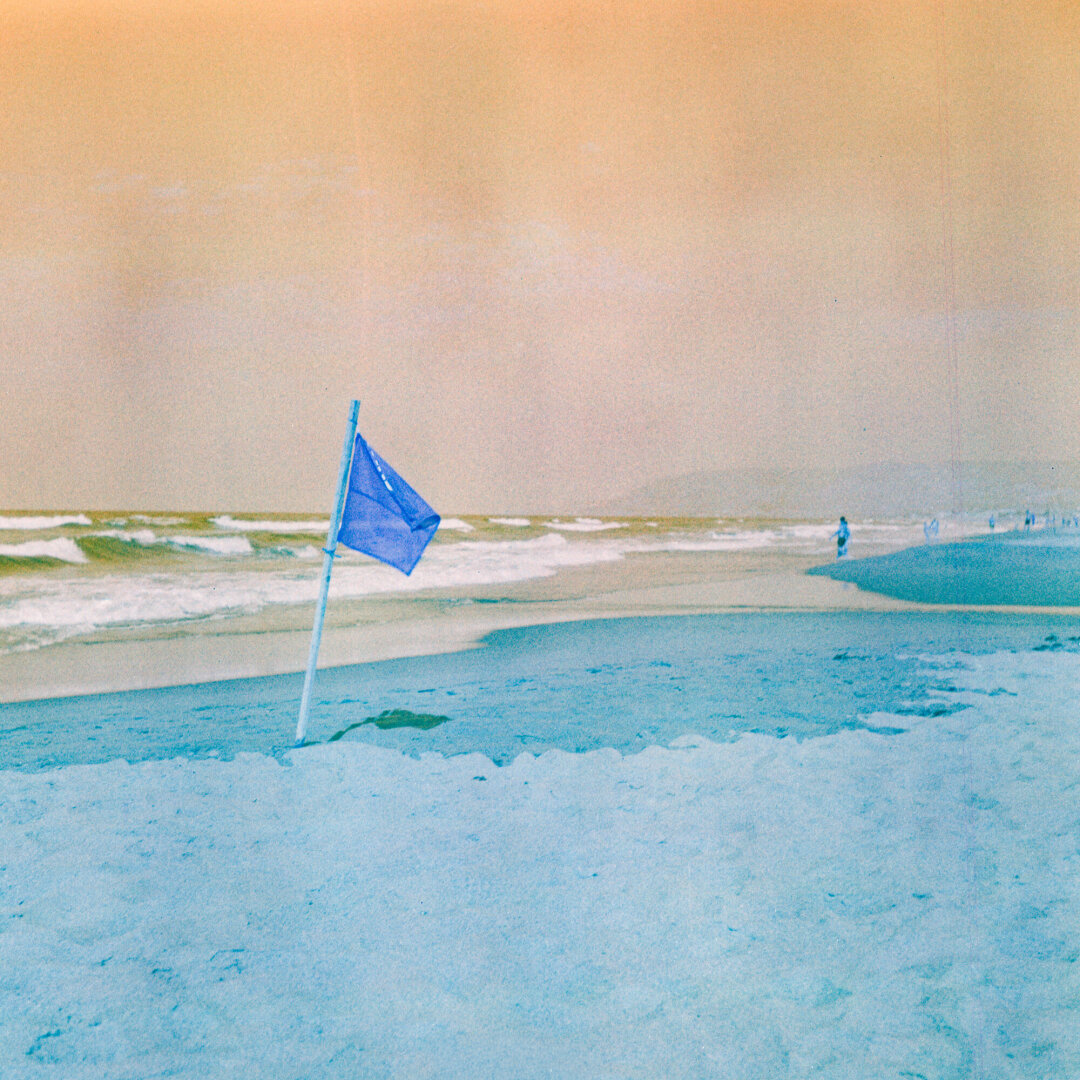 Color photo of a blue flag on a pole  in the sand near the shoreline. Waves roll onto the beach, with a few people visible in the distance. The colors are off: the sky is yellow and the sand and people are blue.