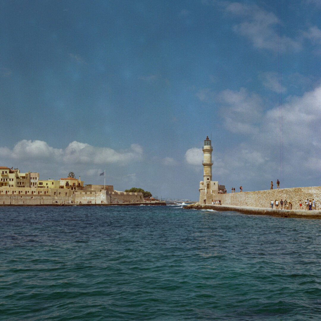 Color photo of a historic stone harbor entrance, with a tall cylindrical lighthouse on the right and low, weathered buildings and fortifications on the left. Deep blue-green water fills the foreground beneath a bright blue sky with scattered white clouds, while small groups of people walk along the stone seawall.