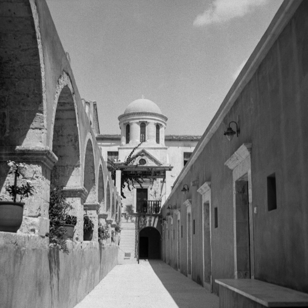 B/W photo of a narrow sunlit passage between stone buildings, with a row of large arches and potted plants on the left. At the far end is a domed classical building with columns and a small balcony, while the right wall has several doorways and wall-mounted lamps.