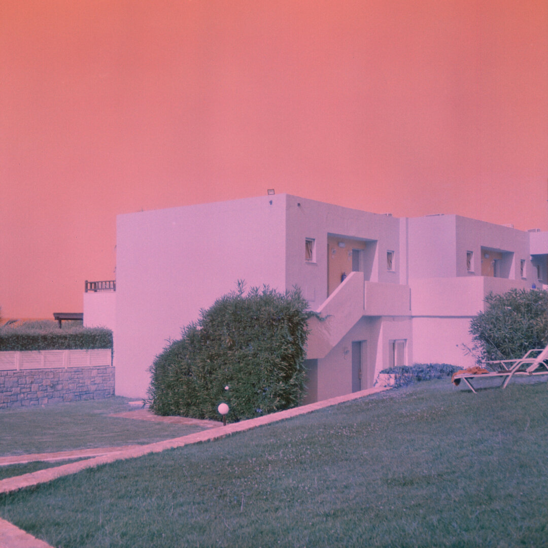 Color photograph of a low-rise resort building surrounded by manicured lawns, shrubs, and lounge chairs. The colors are off. A hazy coral sky and a pink color cast give the quiet scene a dreamy, nostalgic atmosphere.