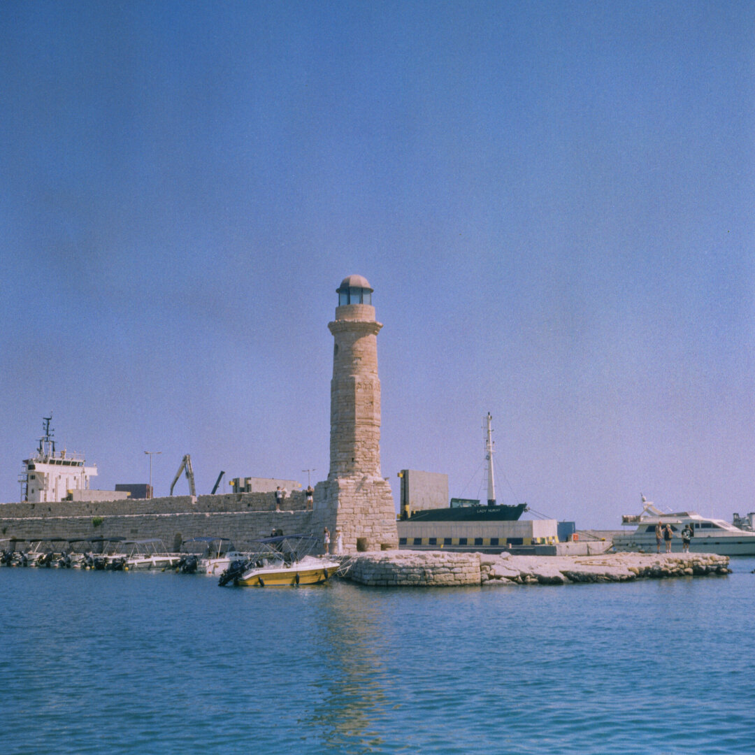 Color photo of a stone lighthouse standing on a small breakwater in calm blue water under a clear blue sky. Several boats, harbor buildings, and a stone seawall surround the lighthouse.