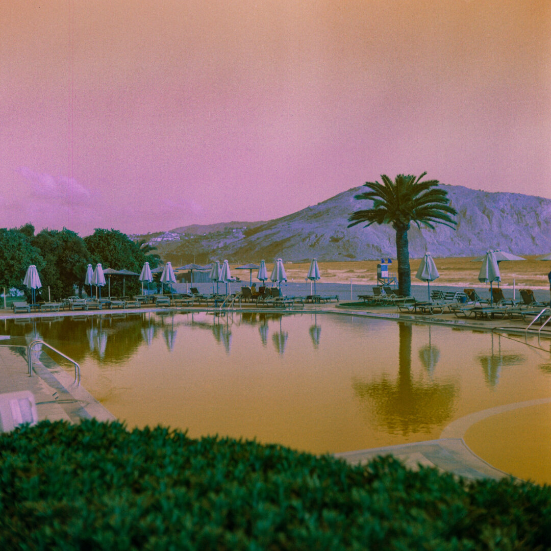 Color photo of a resort pool surrounded by loungers and umbrellas, with a palm tree and rocky mountain in the background. Unusual pink, purple, and greenish tones are characteristic othe experimental film’s color shifts.