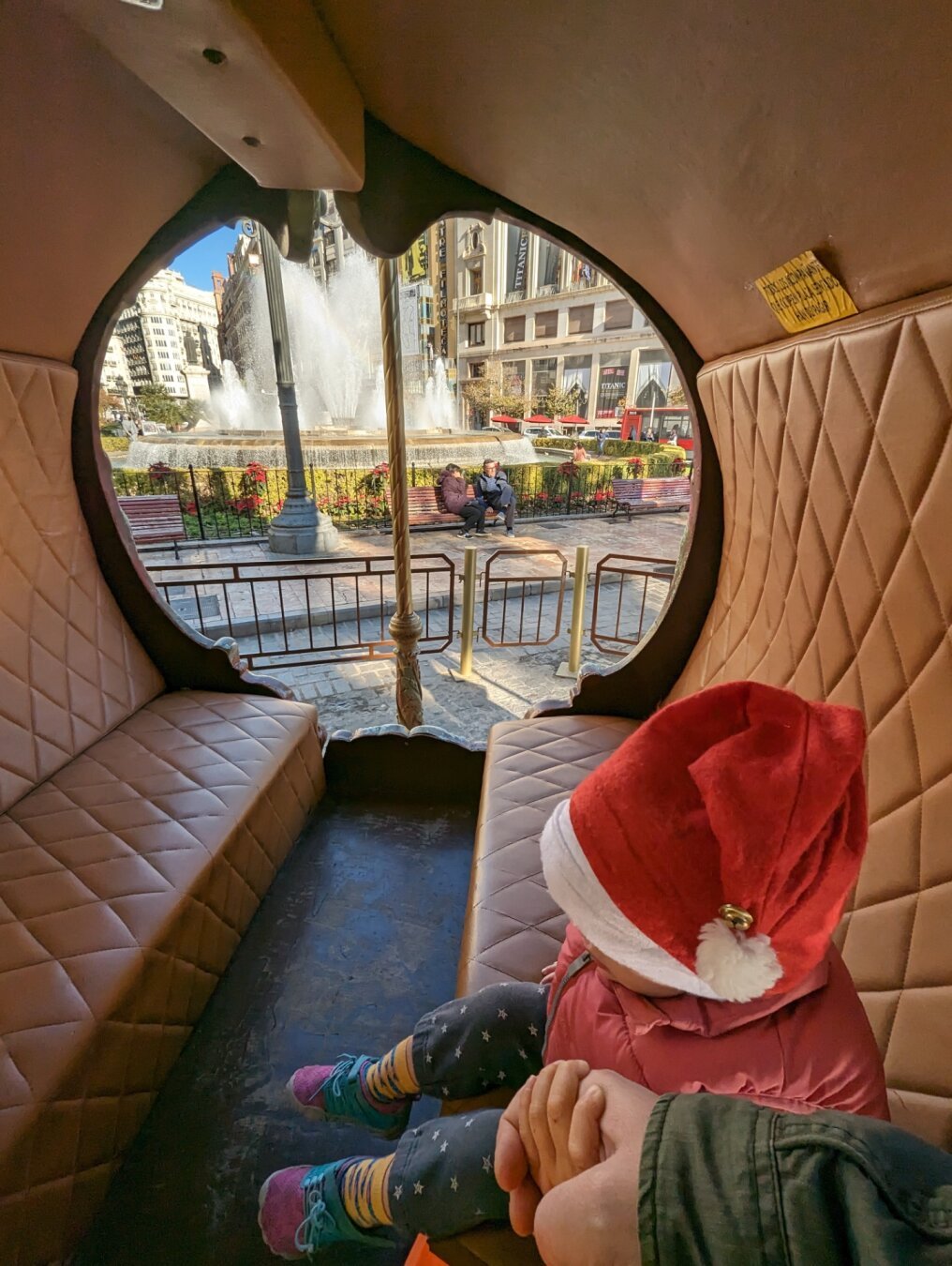 My kid with a Santa hat and holding my hand from a carriage inside a carrousel with a fountain in the back.