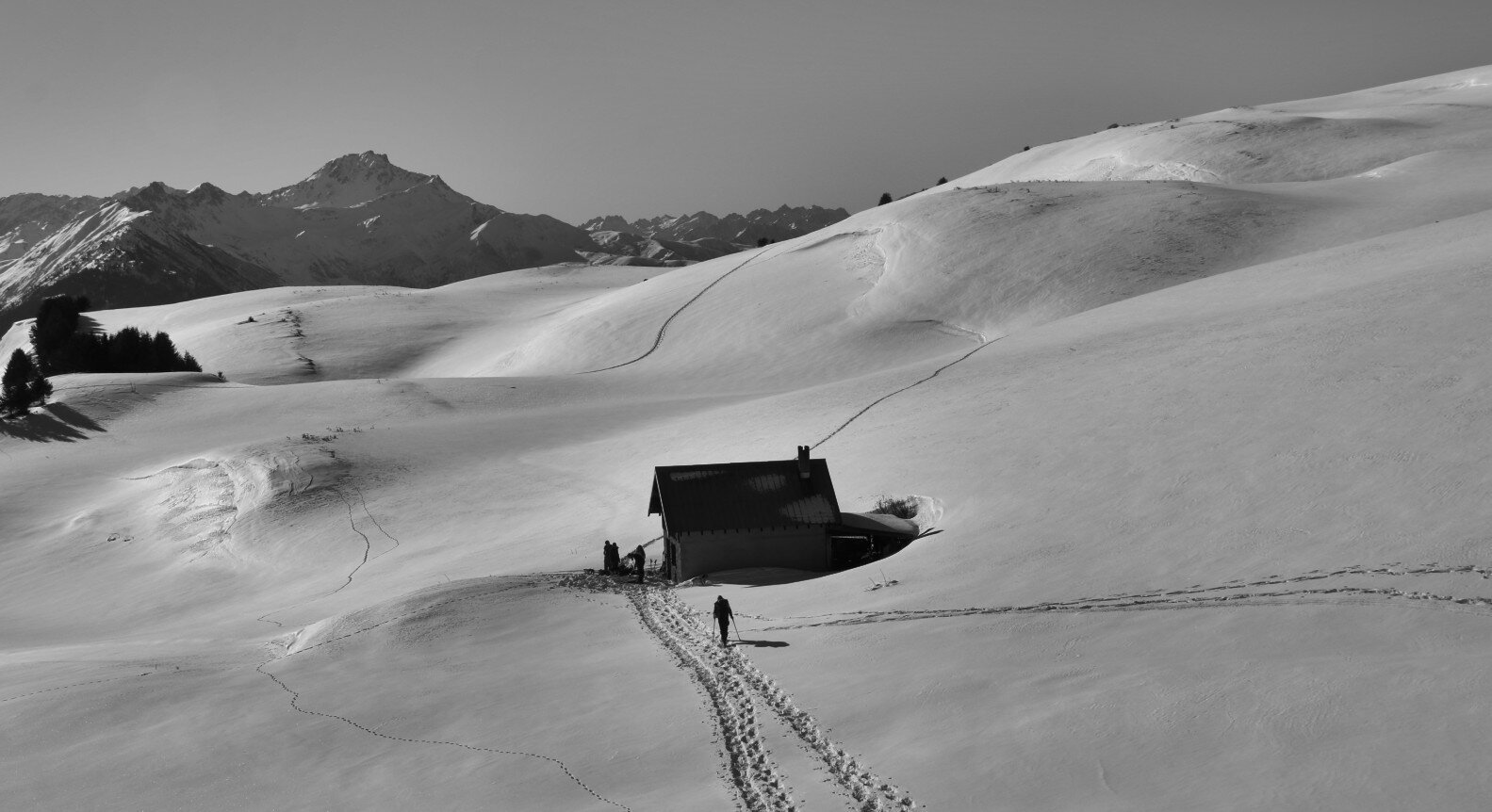 A black and white photo of an unguarded shelter lost on a snowy landscape. We can see people walking around the hut, and the shadows caused by the curves of the snow are visible.