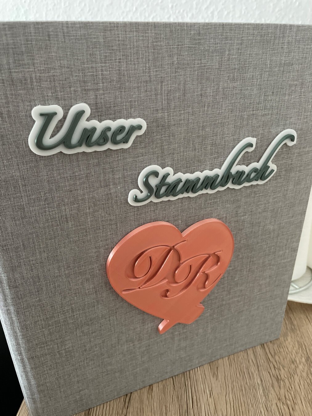 Grey folder with 3 3D prints glued to it. The first one on the upper left corner is „Unser“, the second one in the middle right is „Stammbuch“ and the last one is on the bottom half, it is a rosé heart with embedded characters „D“ and „R“.