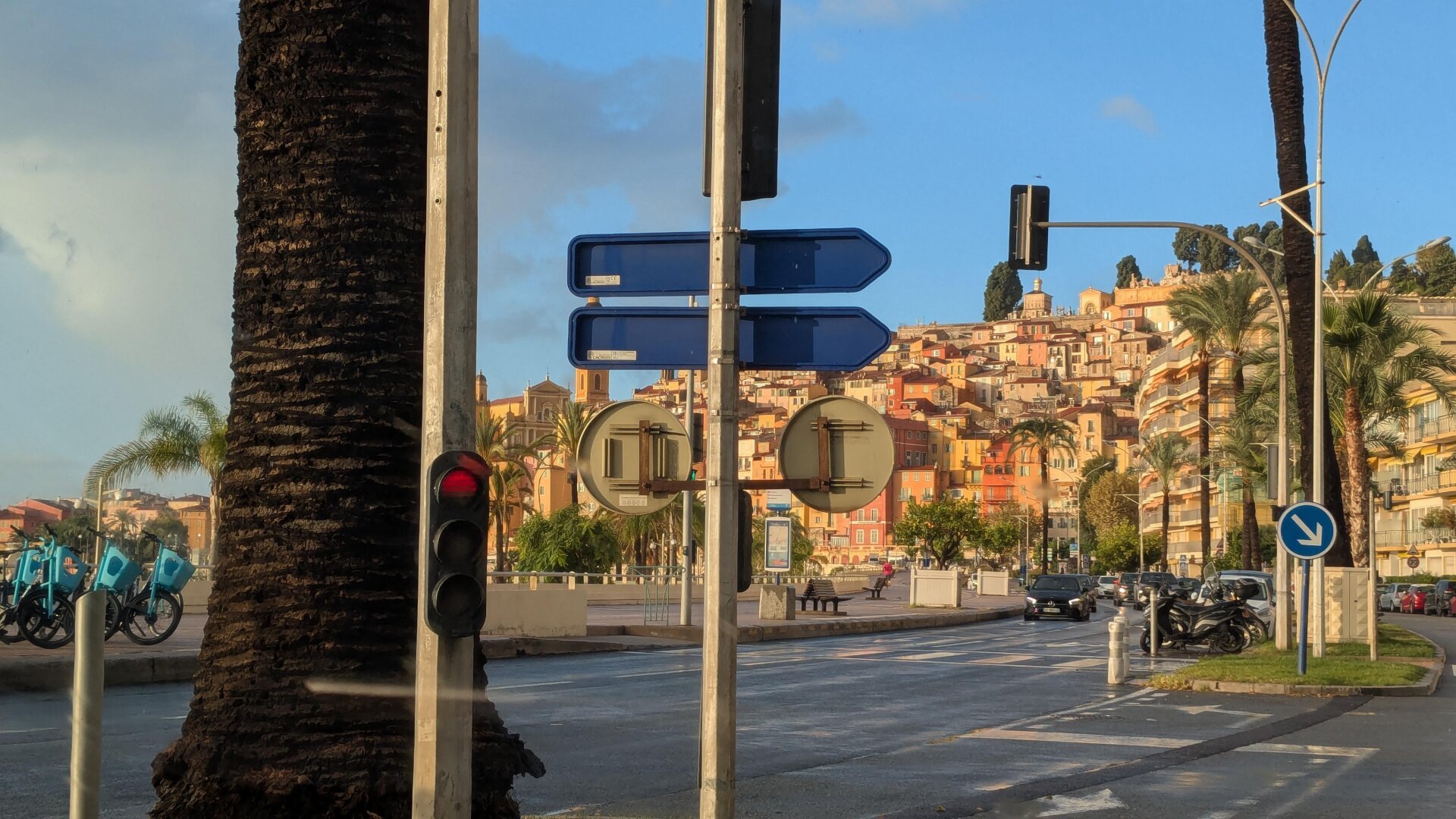 an image from my Pixelfed (because, fuck instagram), shows the city of Menton in the morning sun, except that street signs and palm trees are blocking the view