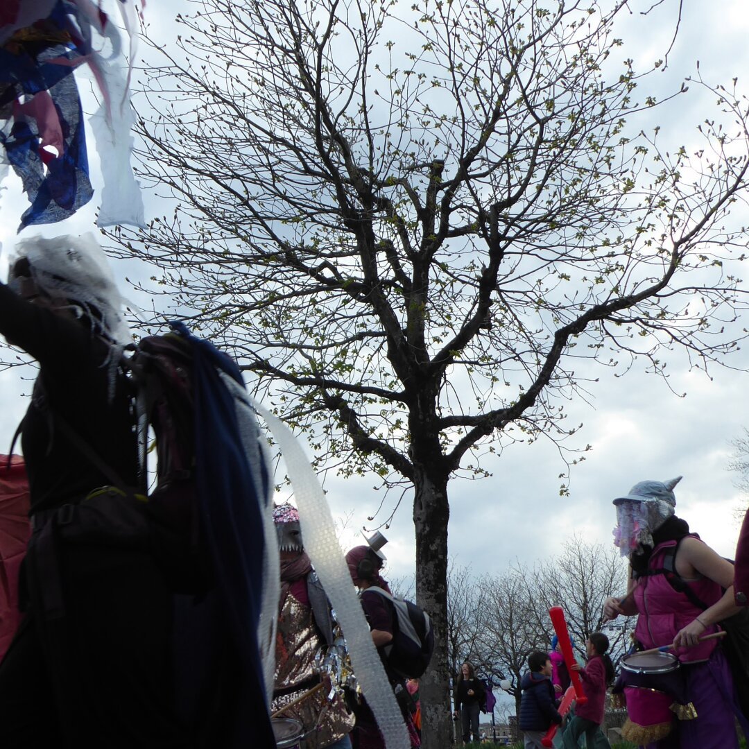 Various masked people are passing the image laterally, some are beating a drum, a large looming figure of a Cthulhu in the left top corner. In the background a tree and some spectators