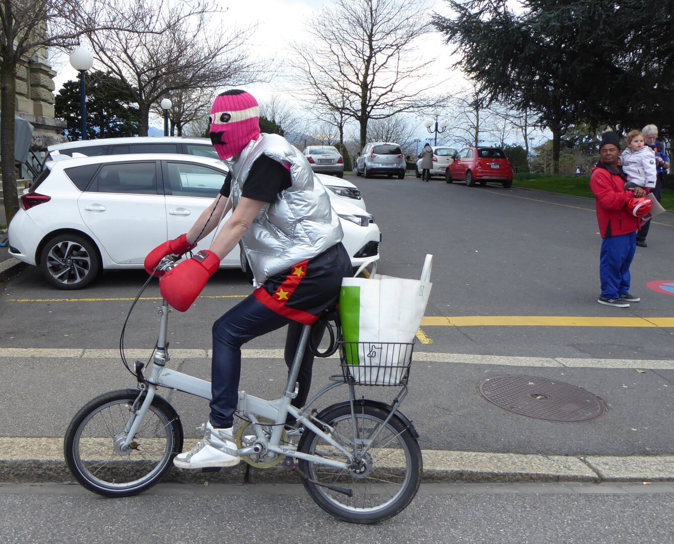 A person sits on a grey bike wearing a pink lucha libre mask and boxing gloves. To the right an older man is looking on while holding a child