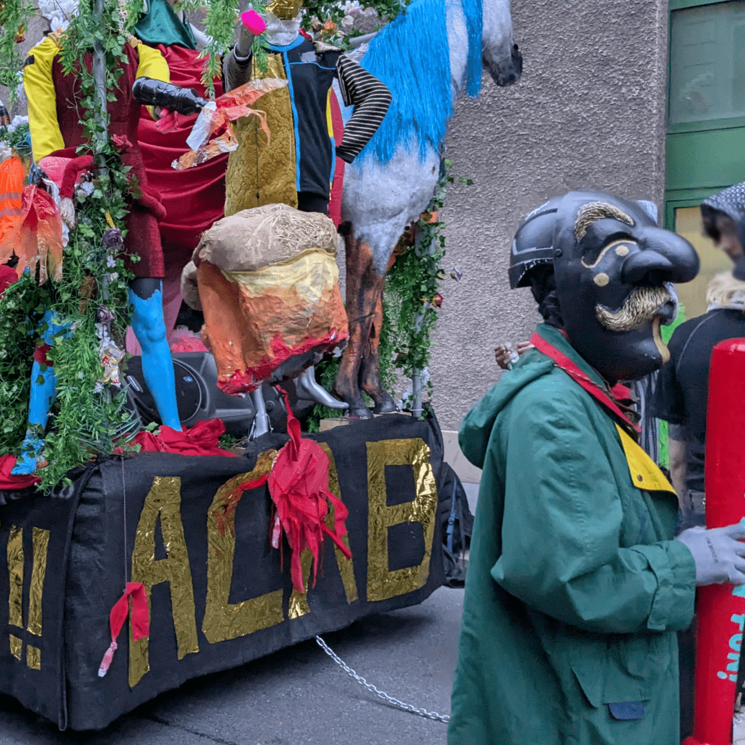 A wildly decorated float in mostly green has the writing ACAB in gold on it, a figure with a big nosed mask stands to the right