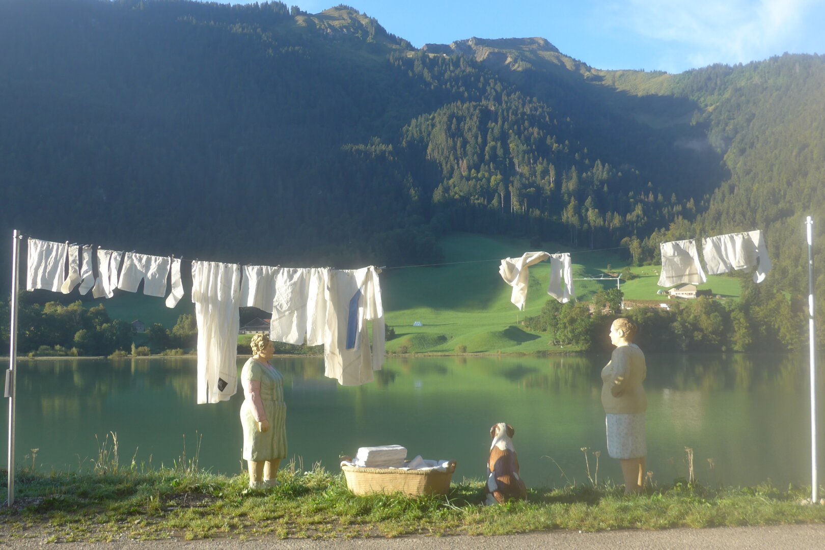an image or two from my Pixelfed, shows a sculpture of a laundry line with two women and a dog near a mountain lake