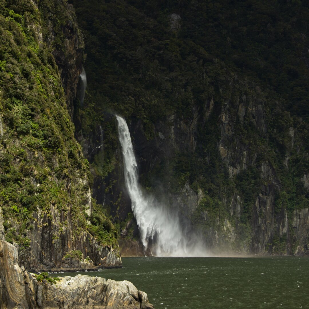 Photo of a waterfall plunging into a fiord, taken from the side of the falls. Wind carries the spray water so that no all of it reaches the bottom.