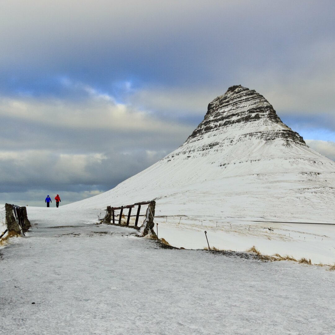 English: picture of Icelandic Kirkjufell mountain to the right. To the left in the foreground is a wooden bridge and two people a bit further along the path. Everything is covered in snow.

Deutsch: Der isländische Berg Kirkjufell rechts im Bild, links im Vordergrund eine hölzerne Brücke, dahinter etwas den Weg entlang spazieren zwei Personen. Alles ist mit Schnee bedeckt.